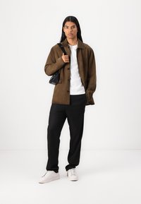 Jachetă din suede maro, tricou alb, pantaloni negri și adidași albi. Geantă mică neagră. Modelul stă în fața unui fundal alb.