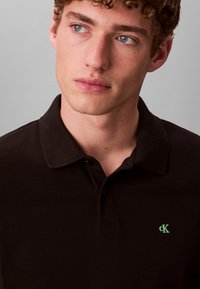 Μαύρο πόλο πουκάμισο από βαμβάκι με κλασικό γιακά, με πράσινο λογότυπο CK. Κοντινή λήψη, που αναδεικνύει την υφή του υφάσματος και τις λεπτομέρειες του κουμπιού.