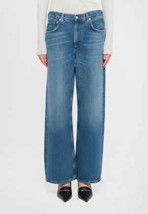 Mujer que lleva pantalones vaqueros de cintura alta y pierna ancha de color azul, combinados con zapatos negros de punta afilada y una blusa de manga larga gris claro.