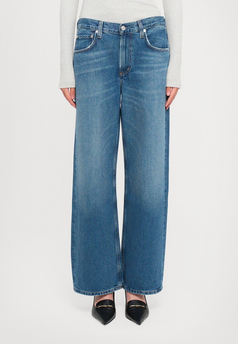 Woman wearing high-waist, wide-leg blue jeans paired with black pointed-toe shoes and a light gray long-sleeve top.