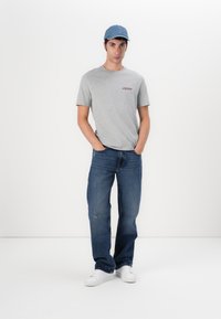 Tricou gri cu mânecă scurtă, având un mic logo roșu, asociat cu blugi din denim bleu. Modelul poartă o șapcă albastră și adidași albi.