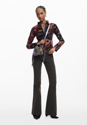 Woman wearing a multicolored patterned shirt, dark flared pants, black pointed shoes, and holding a small colorful crossbody bag.
