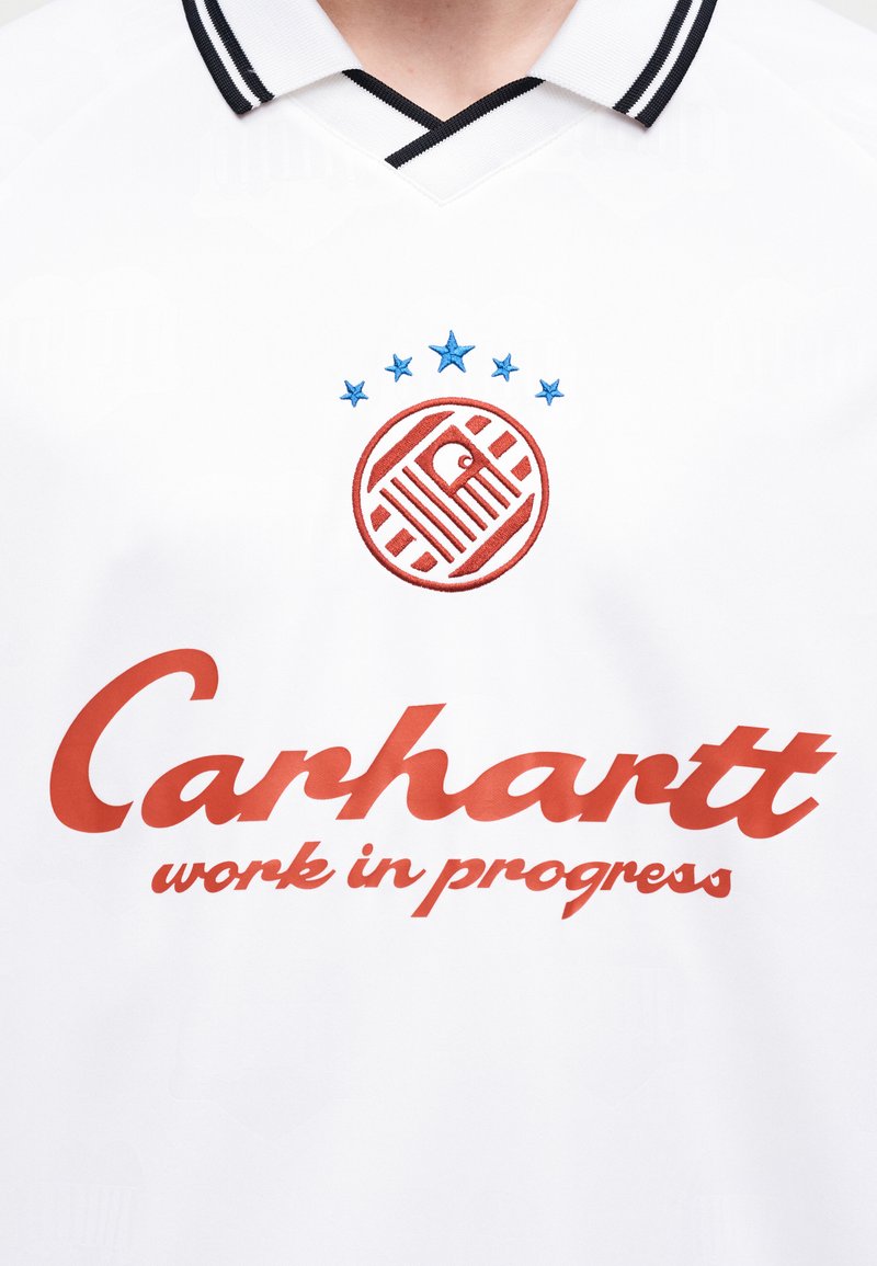 Weißes Poloshirt mit schwarz geringeltem Kragen, darauf ein rotes rundes Emblem mit blauen Sternen und rotem Text „Carhartt work in progress“ darunter.