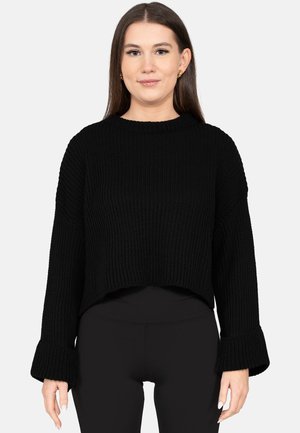 Junge Frau mit langen braunen Haaren, die einen schwarzen, gerippten Strickpullover mit breiten Bündchen und schwarze, eng anliegende Hose trägt, steht vor einem weißen Hintergrund.