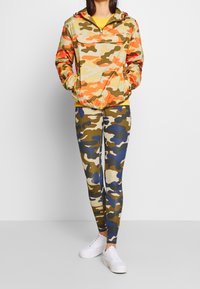 Kamouflage-mönstrad vindjacka i nyanser av orange, beige och grönt, ihop med matchande leggings i blått, brunt och beige kamouflage. Vita sneakers.