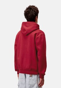 Sudadera roja con capucha, fabricada en un tejido suave, que presenta un corte relajado, puños y dobladillo acanalados, y un bolsillo tipo canguro en la parte frontal.