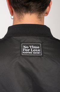 Black jacket featuring a knit collar and a rectangular patch with white text that reads "No Time For Love Kaotiko Society." Smooth texture.
