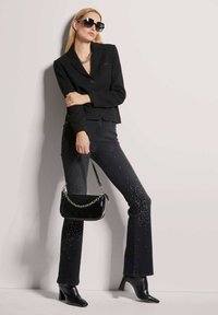 Schwarze, kurze Blazerjacke, schwarze Jeans mit Strassakzenten, schwarze Handtasche aus Lackleder und schwarze Stiefeletten mit Absatz, vor einem neutralen Hintergrund positioniert.