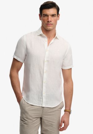 Homme portant une chemise blanche à manches courtes boutonnée et un pantalon beige, debout avec une main dans la poche, sur un fond uni.