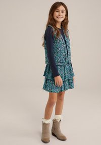 Kvetinové šaty s volánmi v modrej, zelenej a bielej farbe. Viacvrstvový vest, dlhé námornícke rukávy a béžové čižmy s čipkovaným lemom.