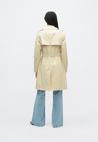 Béžový trenčkot s opaskom v páse a zapínanou klopou na zadnej strane, nosený cez svetlomodré rozšírené džínsy. Hladká textúra, minimalistický dizajn.