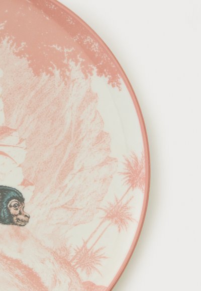 Assiette ronde en céramique avec un motif tropical rose et blanc présentant des palmiers et un visage de singe près du bord.