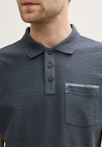 Polo gris foncé en tissu texturé, avec trois boutons et une poche poitrine ornée d'une bande bleu clair.