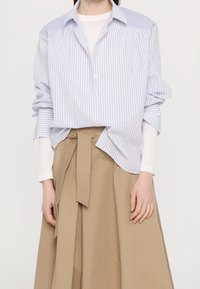 Nainen, jolla on päällään vaaleansininen ja valkoinen raidallinen napillinen paita valkoisen pitkähihaisen paidan päällä sekä korkeavyötäröinen beige hame, jossa on solmittu vyö.