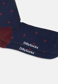 Chaussettes navy avec des pois rouges, présentant un accent bordeaux au niveau du talon. Logo "DillySocks" imprimé en blanc sur le bord. Texture côtelée.