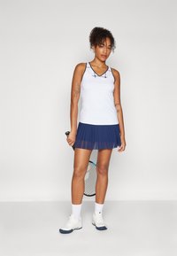 Top blanco sin mangas con una falda plisada de color marino, que presenta un patrón de cuadros en el escote. La modelo sostiene una raqueta de tenis y lleva calzado blanco.