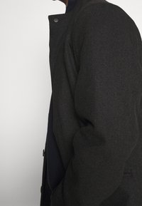 Cappotto scuro in lana con una texture liscia, caratterizzato da un colletto alto e chiusure a scatto. Le maniche sono leggermente oversize, offrendo una vestibilità comoda.