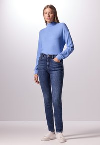 Suéter de cuello alto azul claro, de textura suave; jeans ajustados azul oscuro; zapatillas blancas; modelo con las manos en los bolsillos, contra un fondo claro.