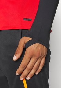 Rode sportshirt met zwarte lange mouwen, voorzien van een duimopening. Zwarte broek met een verticale oranje accent. JAKO-logo zichtbaar.