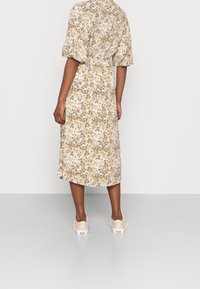Robe midi fleurie en tons crème, marron et or, avec des manches bouffantes et une taille élastique. Des chaussures décontractées beige clair complètent la tenue.