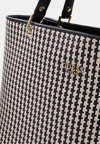 Sac fourre-tout tissé noir et blanc avec des poignées en cuir noir et le logo doré "GUESS EST 1981" à l'avant.