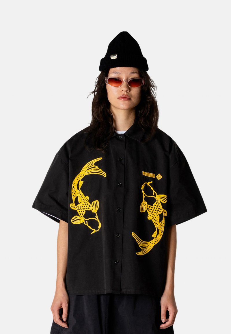 Femme portant un bonnet noir, des lunettes de soleil translucides rouges et une chemise noire à manches courtes avec de grands motifs de poissons koi jaunes.
