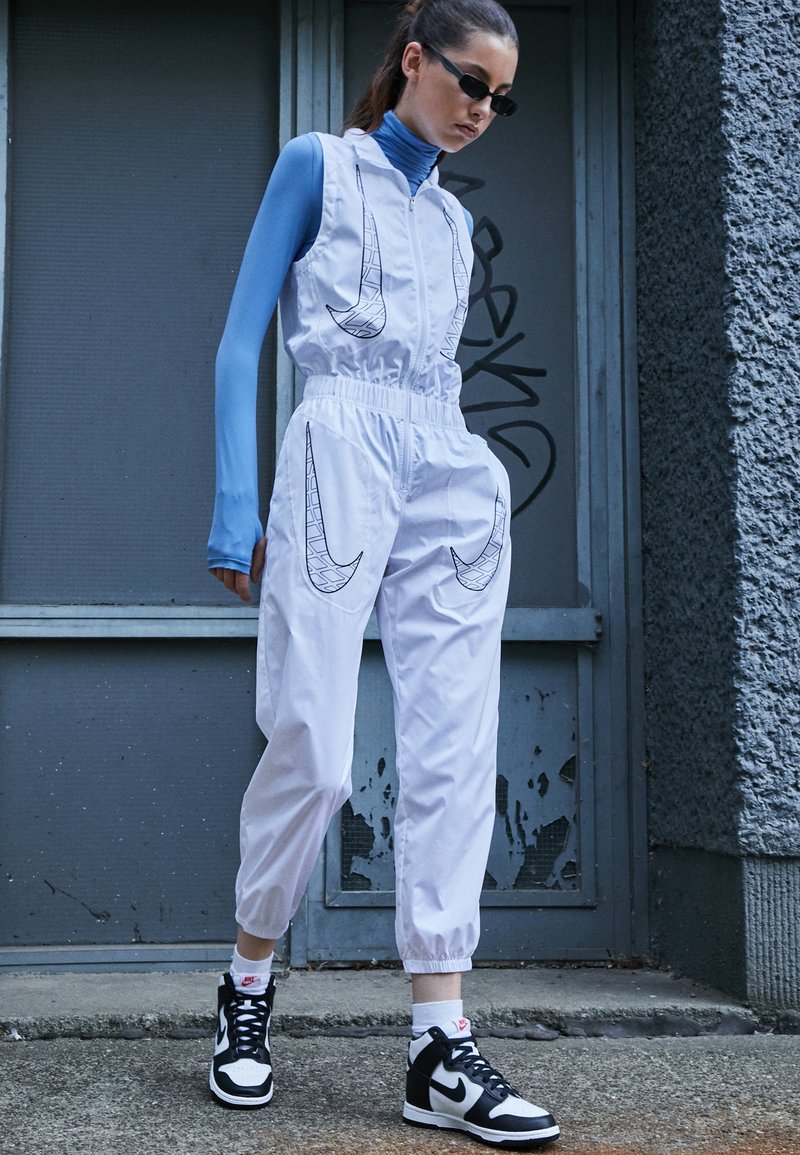 Mono blanco con gráficos de swoosh negros, cinturilla elástica y patas acampanadas. Llevado sobre una camiseta de manga larga azul y zapatillas negras.
