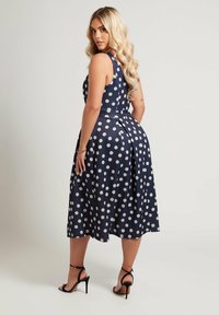 Vestido de lunares azul marino con un corsé ajustado y falda acampanada, que presenta un detalle de pliegue en la espalda. Usado con tacones negros de tiras.