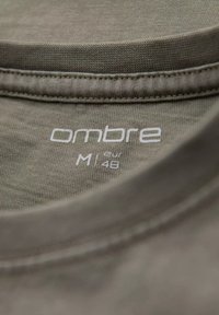 Šedá bavlněná tričkovina s kulatým výstřihem, s etiketou, na které je bílým písmem napsáno "ombre M eur 48". Hladká textura a jednoduchý design.