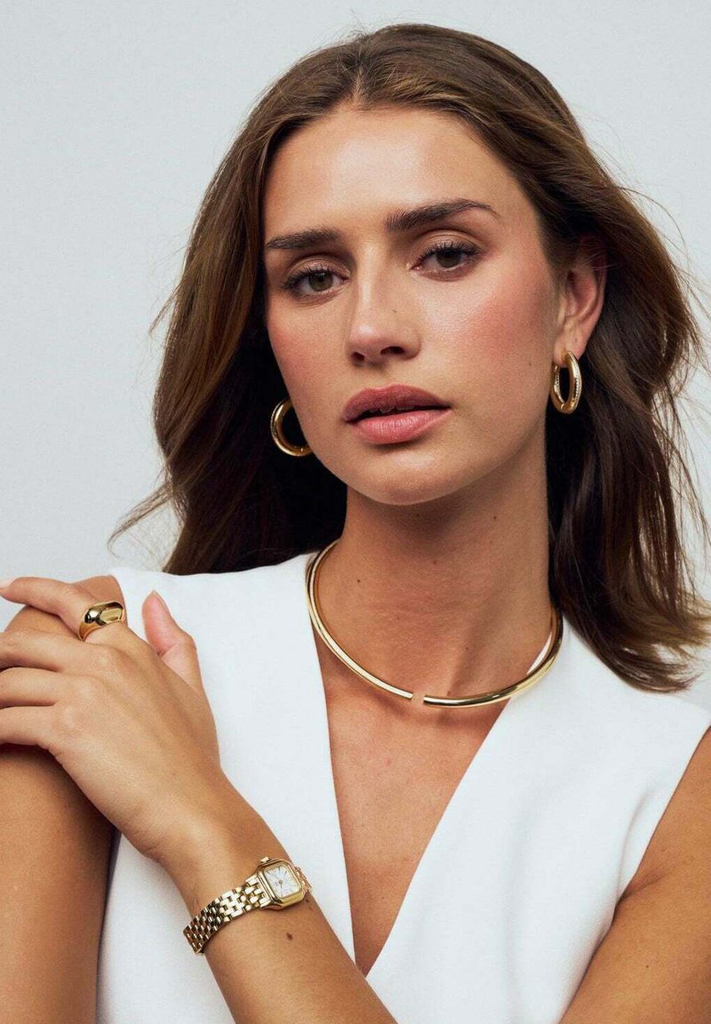 Guldtonad smycken som inkluderar en kraftig choker, hoopörhängen och en minimalistisk klocka med fyrkantig urtavla och metallarmband. Släta texturer med metallisk glans.