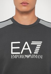 Gros plan sur une personne portant un t-shirt gris foncé EA7 Emporio Armani avec des rayures blanches sur les épaules et le logo blanc sur la poitrine.