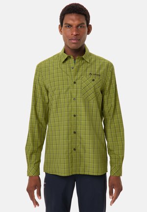 Uomo che indossa una camicia verde a quadri a maniche lunghe con taschino frontale e pantaloni scuri, in piedi di fronte.