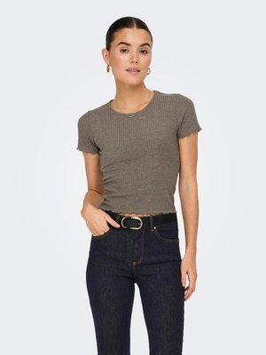 Kvinne med mørkt hår i en knute, iført en kortermet, ribbet taupe topp og mørke høytlivsjeans med svart belte, hånden i lommen.