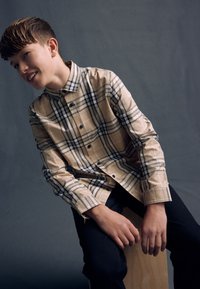 Beige kariertes Hemd mit blauen und schwarzen Akzenten, zugeknöpft, kombiniert mit dunklen Jeans. Person sitzt auf einem hölzernen Hocker vor einem grauen Hintergrund.