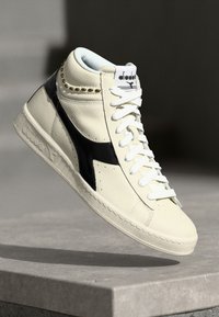 High-top sneaker featuring cream leather, black accents, gold studs, and white laces. Textured sole and contrast stitching present.