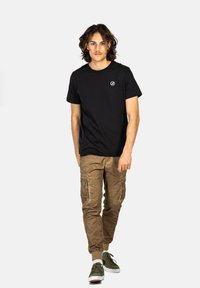 Schwarzes Baumwoll-T-Shirt mit kurzen Ärmeln und Logo auf der Brust, kombiniert mit braunen Cargo-Hosen, die über Seitentaschen und elastische Bündchen verfügen. Grüne Sneakers.