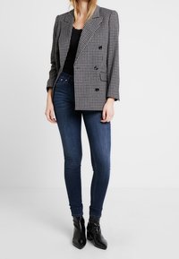 Frau in schwarzem, eng anliegendem Oberteil, grauem karierten, doppelt knöpfbaren Blazer, dunklen Skinny-Jeans und schwarzen spitzen Stiefeletten vor weißem Hintergrund.