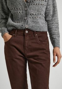 Grå stickad cardigan med knappfront och öppen väv, i kombination med bruna denimbyxor som har en klassisk femfickdesign och metallaccenter.