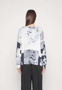 Haut à manches longues en blanc et gris avec des motifs floraux et abstraits ; il possède un col rond et des poignets côtelés ; associé à des pantalons plissés noirs.