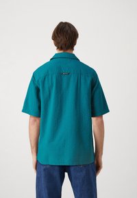 Maglietta a maniche corte color turchese con un tessuto testurizzato, colletto e un sottile etichetta del marchio sulla parte posteriore. Le cuciture dettagliate migliorano il design.