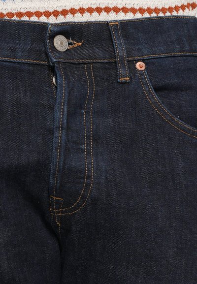 Jeans de mezclilla azul oscuro con herrajes de cobre, detalles de costura visibles y un diseño clásico de cinco bolsillos. La tela tiene una textura sutil.
