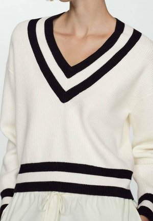 White knit sweater with black double stripes on V-neck, cuffs, and waist, worn over a light-colored garment with a drawstring.