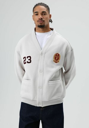 Lichtgrijze cardigan met sjaalkraag, geborduurde patches, knoopsluiting en voorzakken, gedragen over een wit overhemd en donkere spijkerbroek.