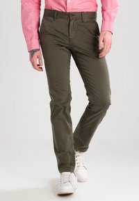 Olivgrüne Chinos mit schmaler Passform, Fünf-Taschen-Design und braunem Knopfverschluss. Kombiniert mit weißen Sneakers.