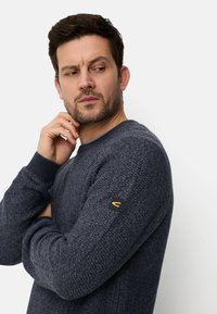 Granatowy sweter dziany z teksturowanym wzorem, ściągaczami na mankietach oraz czarną naszywką z logo na lewym rękawie. Okrągły dekolt i uniwersalny fason.