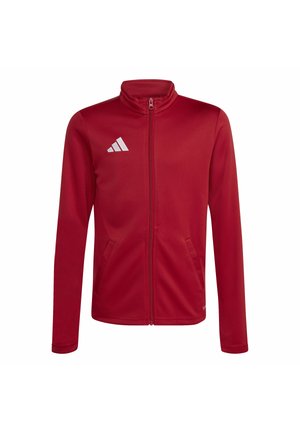 Młody sportowiec ubrany w czerwoną kurtkę na zamek błyskawiczny i czarne spodnie trzyma pod pachą białą i czarną piłkę nożną Adidas.