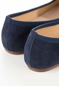 Ballerines en suede bleu marine avec un bout rond, un bord élastiqué et une semelle en caoutchouc brun, présentant des coutures soignées à l'arrière.