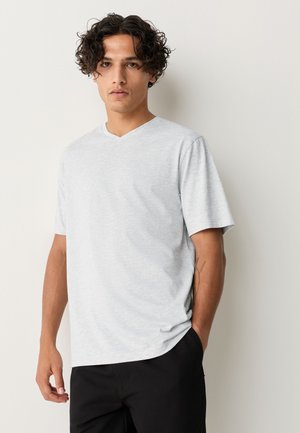 Ung mand med krøllet hår iført en lysegrå T-shirt med korte ærmer og V-udskæring samt sorte bukser, stående op ad en enkel, lys væg.