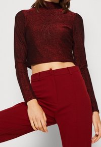 Roter glitzernder, kurz geschnittener Langarmoberteil mit hohem Kragen, kombiniert mit roten, eng anliegenden Hosen, die eine glatte Textur und schimmernde Details zeigen.