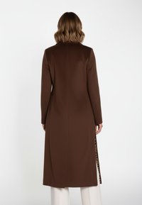 Brun lang frakke med en glat tekstur, set bagfra med et slidsspor med leopardmønsterdetalje, skræddersyet pasform og klassisk krave.